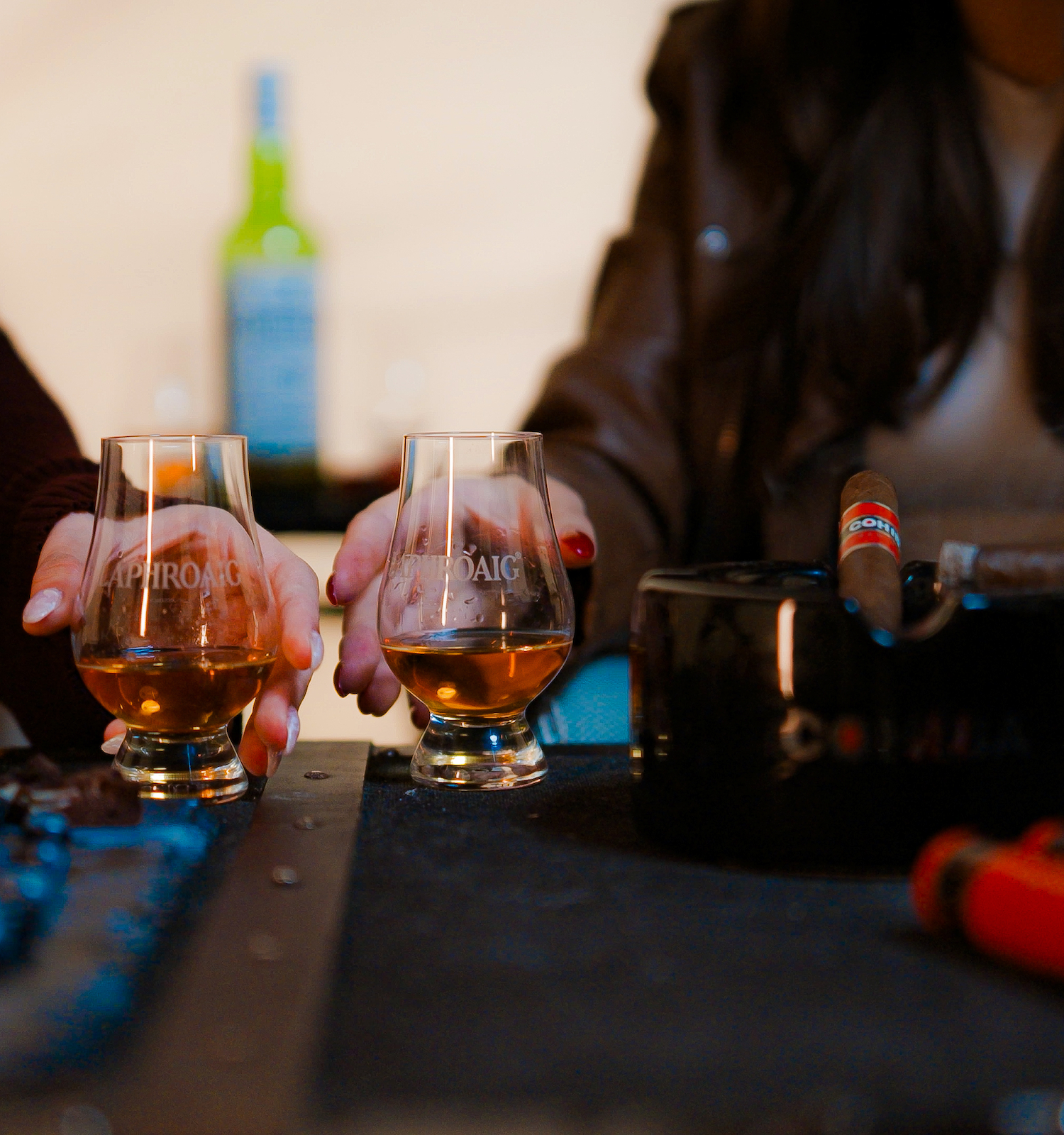 Two people hold glasses of amber liquid by a table with cigars, a bottle, and a cheese platter.