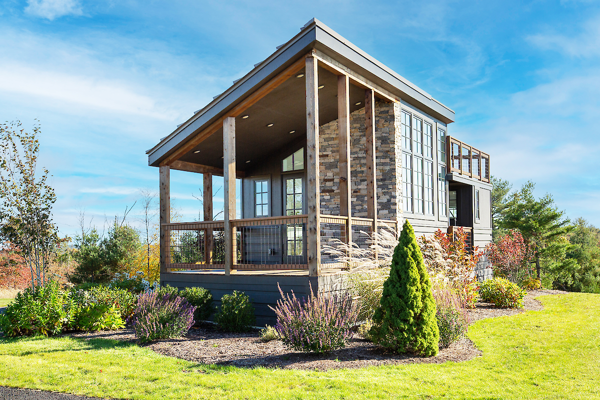 A modern tiny house with large windows and a wooden porch, surrounded by a neatly landscaped garden under a clear blue sky.