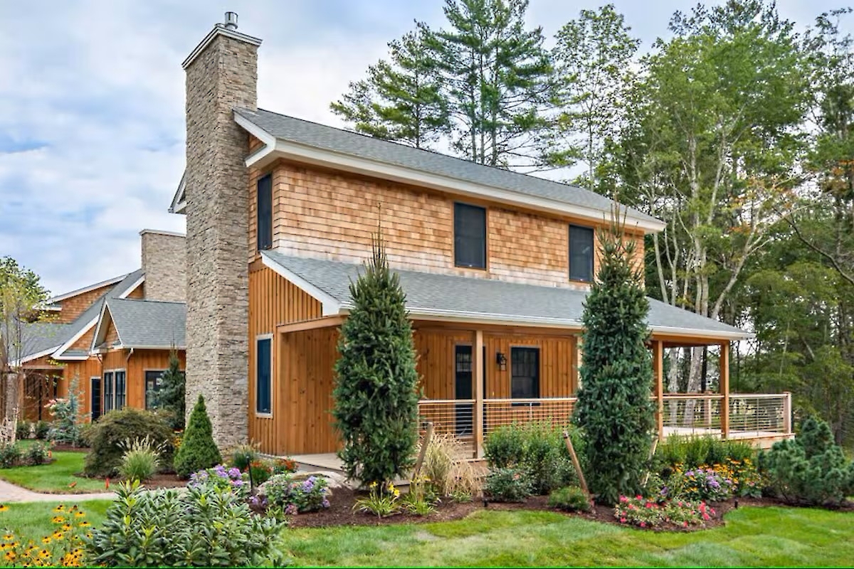 The image shows a rustic house with wood siding, a stone chimney, surrounded by lush greenery and a neatly landscaped garden.