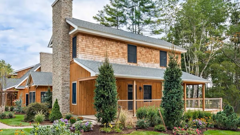 The image shows a rustic house with wood siding, a stone chimney, surrounded by lush greenery and a neatly landscaped garden.