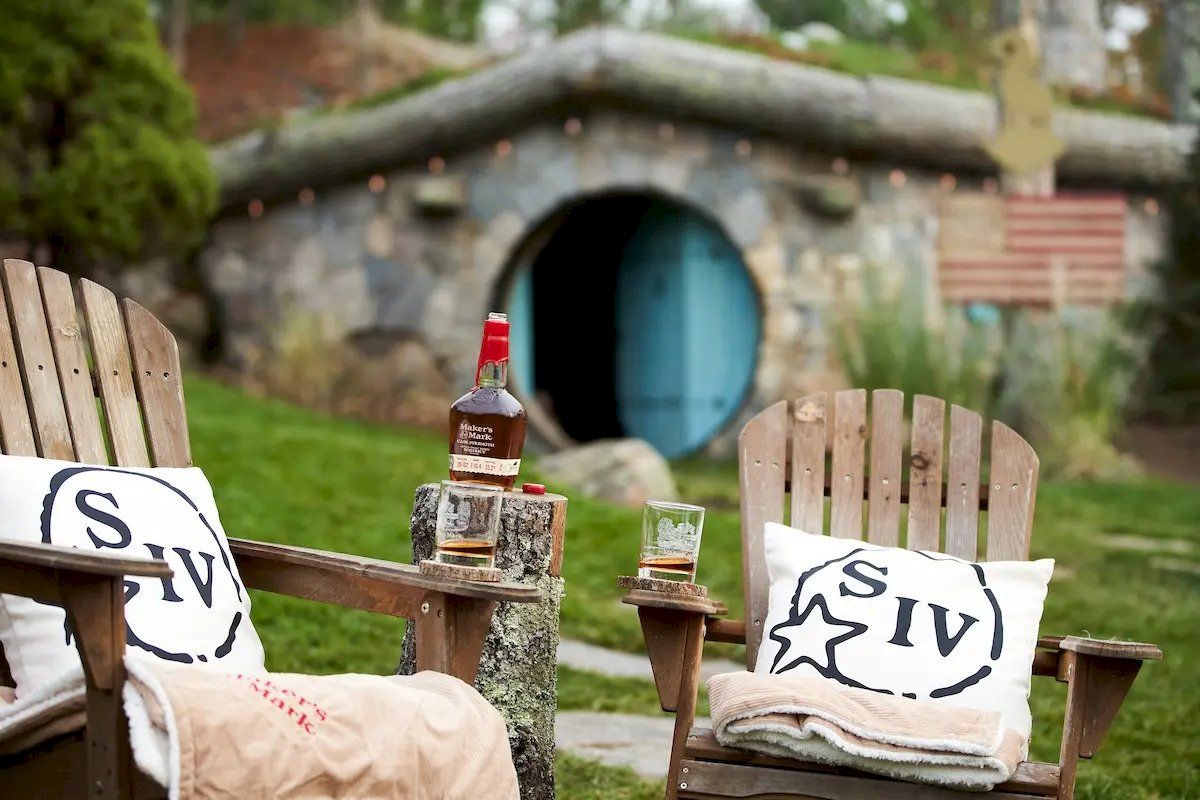 Two wooden chairs with pillows and a small table holding a bottle and two glasses in front of a Hobbit-like house with a blue door.