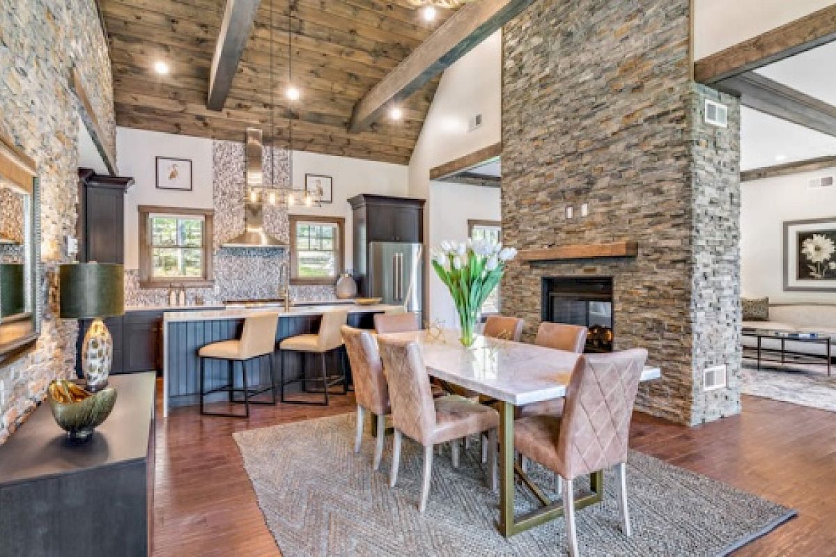 This image shows a modern open-concept kitchen, dining room, and living room with wooden beams, stone walls, and contemporary furnishings.