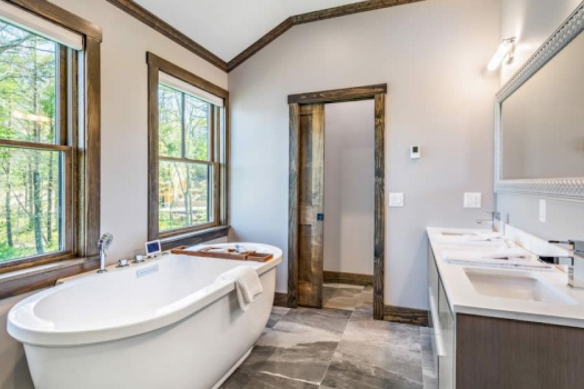 A spacious bathroom with a freestanding bathtub, two windows, double sinks with a large mirror, and wood accents on doorframes and trim.