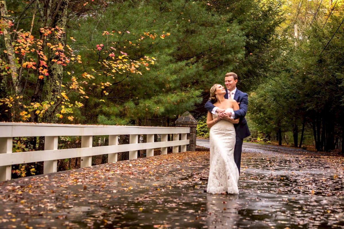 A couple dressed in wedding attire embraces and smiles on a wet road surrounded by autumn foliage, next to a white fence.