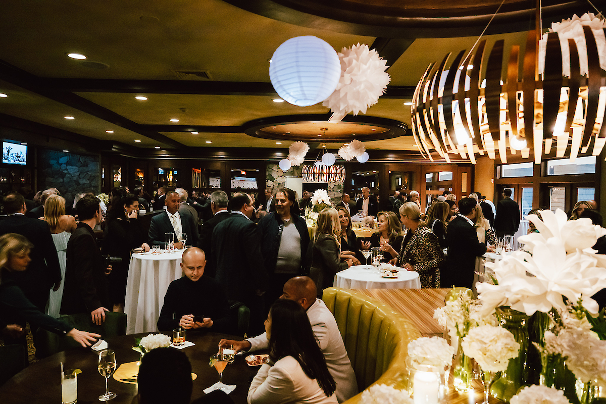 A lively indoor gathering with people socializing around tables, elegant decor, and warm lighting creates a festive atmosphere.