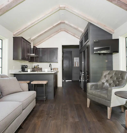 The image shows a cozy living area with a sofa, armchair, and small kitchen, featuring wooden floors and large windows for natural light.