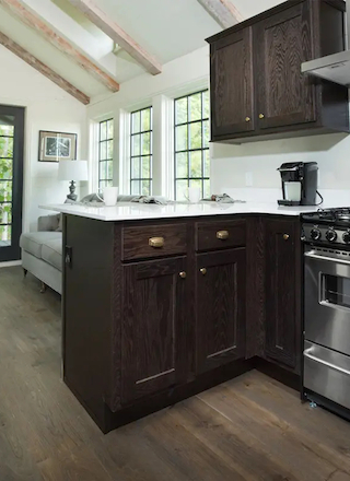 A cozy kitchen with dark cabinets, a stove, and a coffee maker. There's a living area with a sofa and large windows in the background.