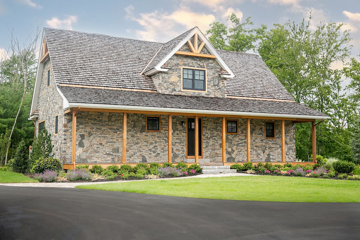 A stone house with a wooden porch and a gabled roof, surrounded by a well-maintained lawn and trees, on a clear day.