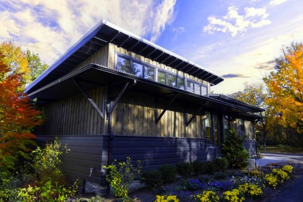 A modern wooden house with large windows, surrounded by greenery and colorful autumn trees under a partly cloudy sky.