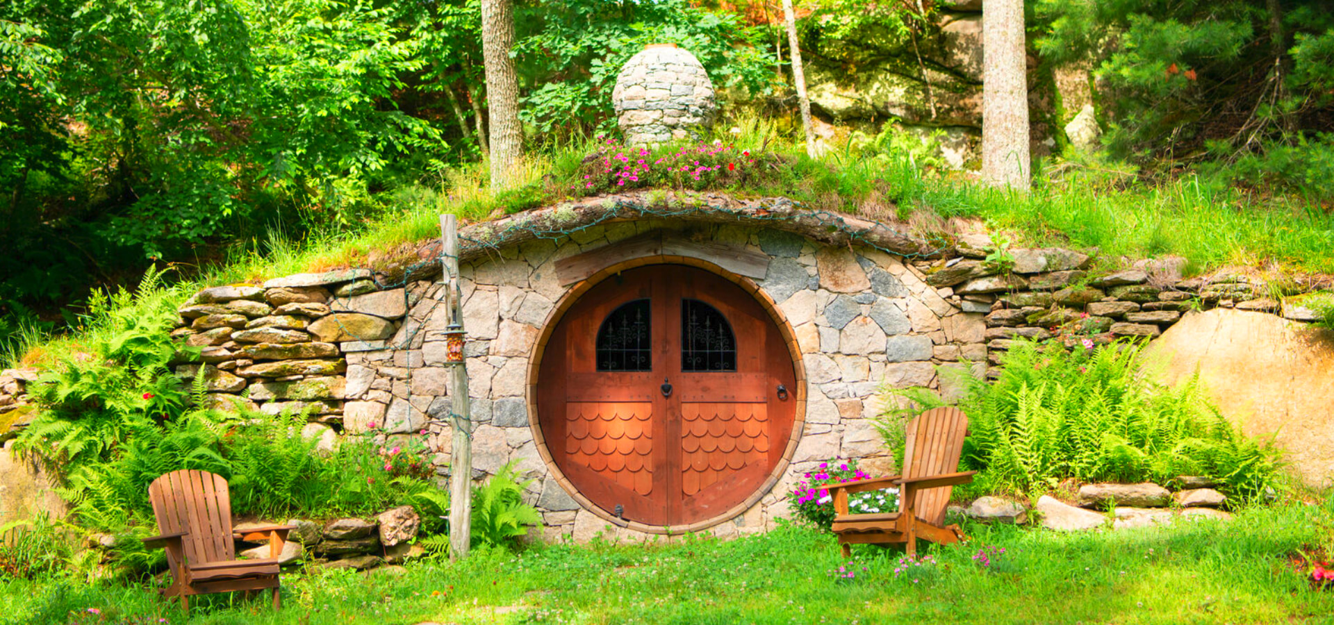 A charming, hobbit-like house with a round wooden door set in a grassy hillside, flanked by two wooden chairs, and lush green surroundings.