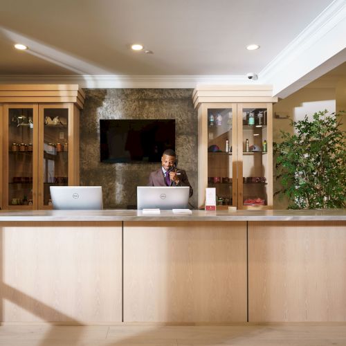 A modern reception desk with a person at the counter, two computers, shelving units, a TV on the wall, and various decor pieces in the background.