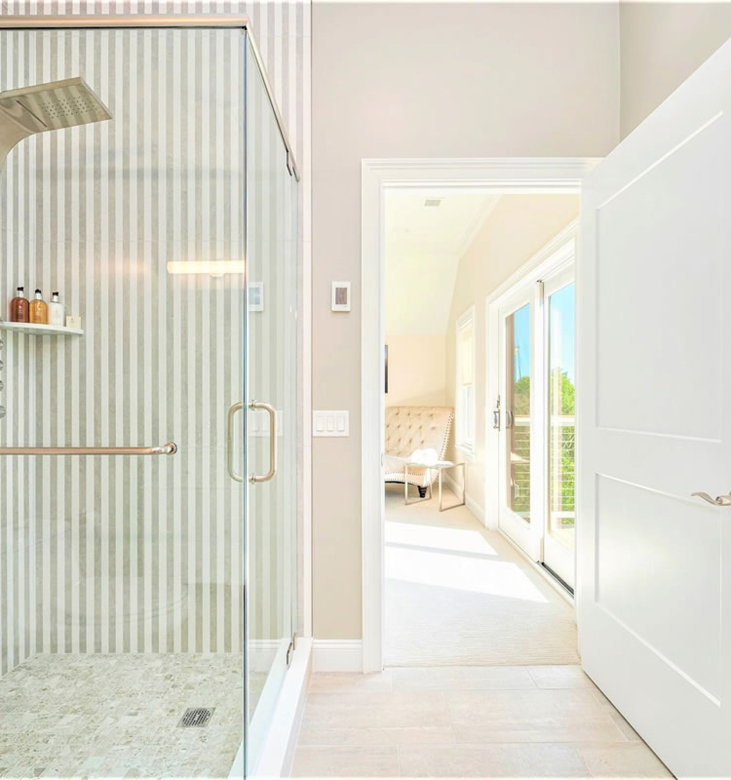 A modern bathroom with a glass-enclosed shower featuring multiple shower heads and shelves. A white door leads to a bright, sunlit room beyond.