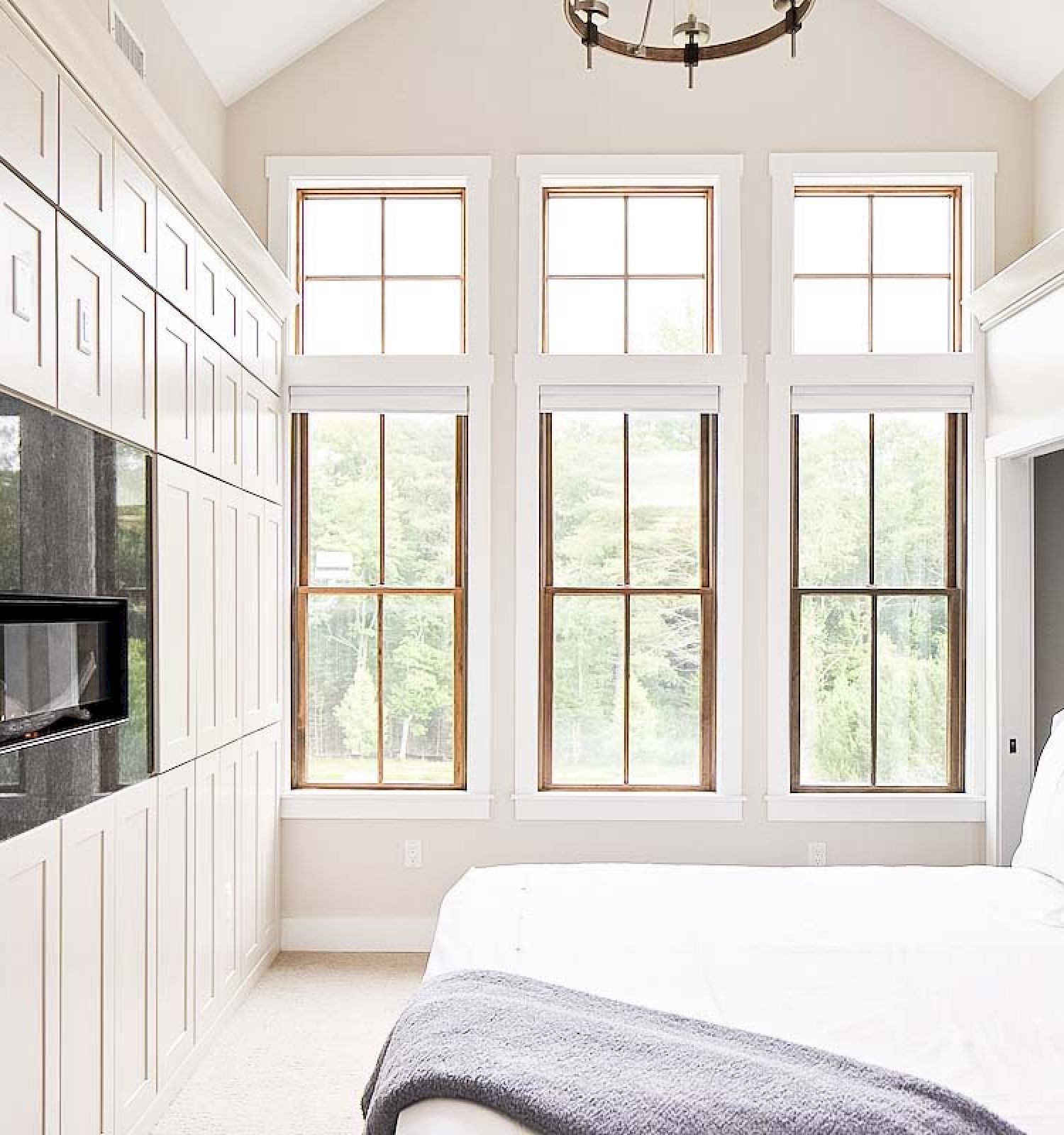 A bright bedroom with tall windows, a white bed, built-in cabinetry, a TV, and a chandelier, all bathed in natural light.