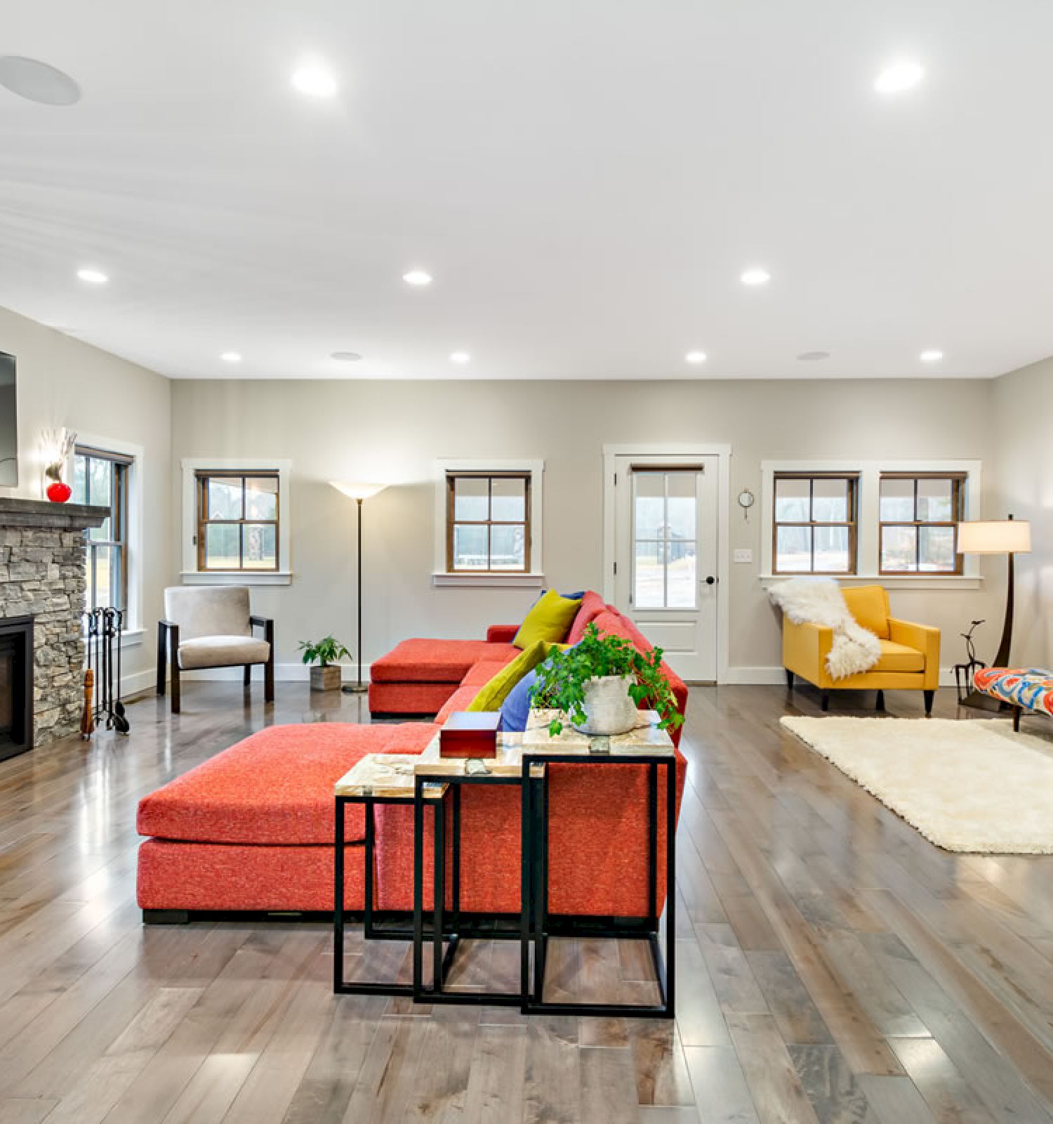 A spacious, modern living room with colorful furniture, a stone fireplace, wall-mounted TV, and large windows letting in natural light.