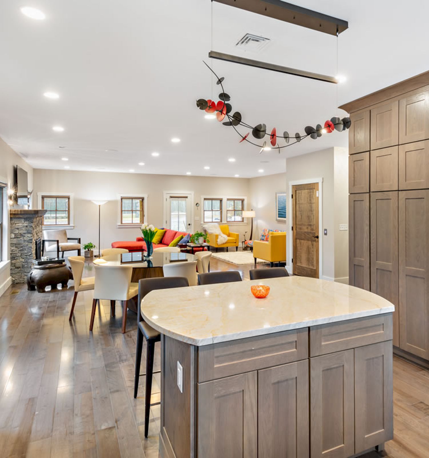 The image shows a modern open-concept kitchen and living area with wood cabinetry, a large island, stainless steel appliances, and colorful seating arrangements.