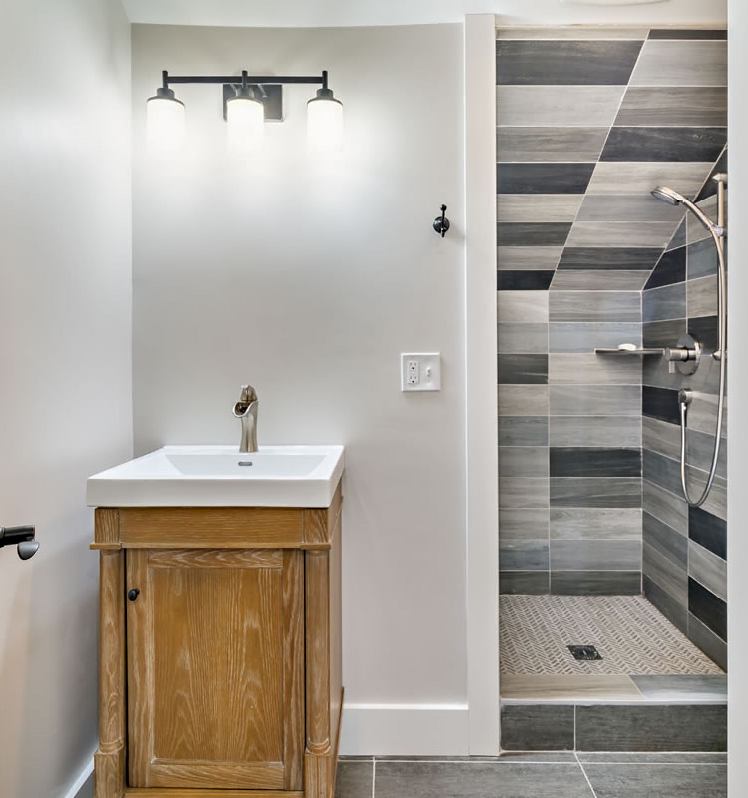 This image shows a modern bathroom with a wooden vanity, sink, and illuminated mirror. There's also a tiled walk-in shower with a glass door ending the sentence.