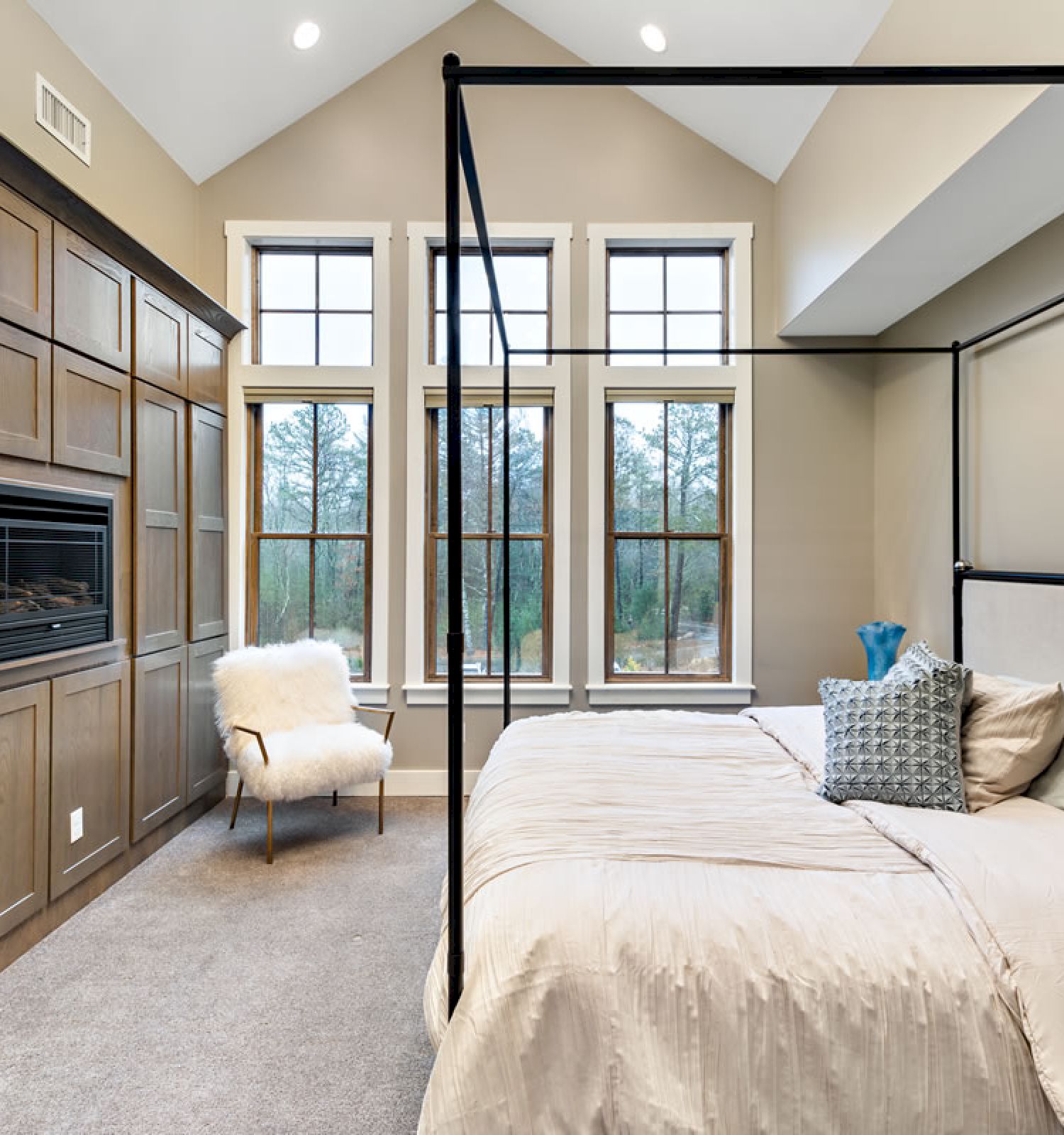 A modern bedroom with a large bed, tall windows, a cozy chair, and built-in wooden cabinets surrounding a TV or fireplace.