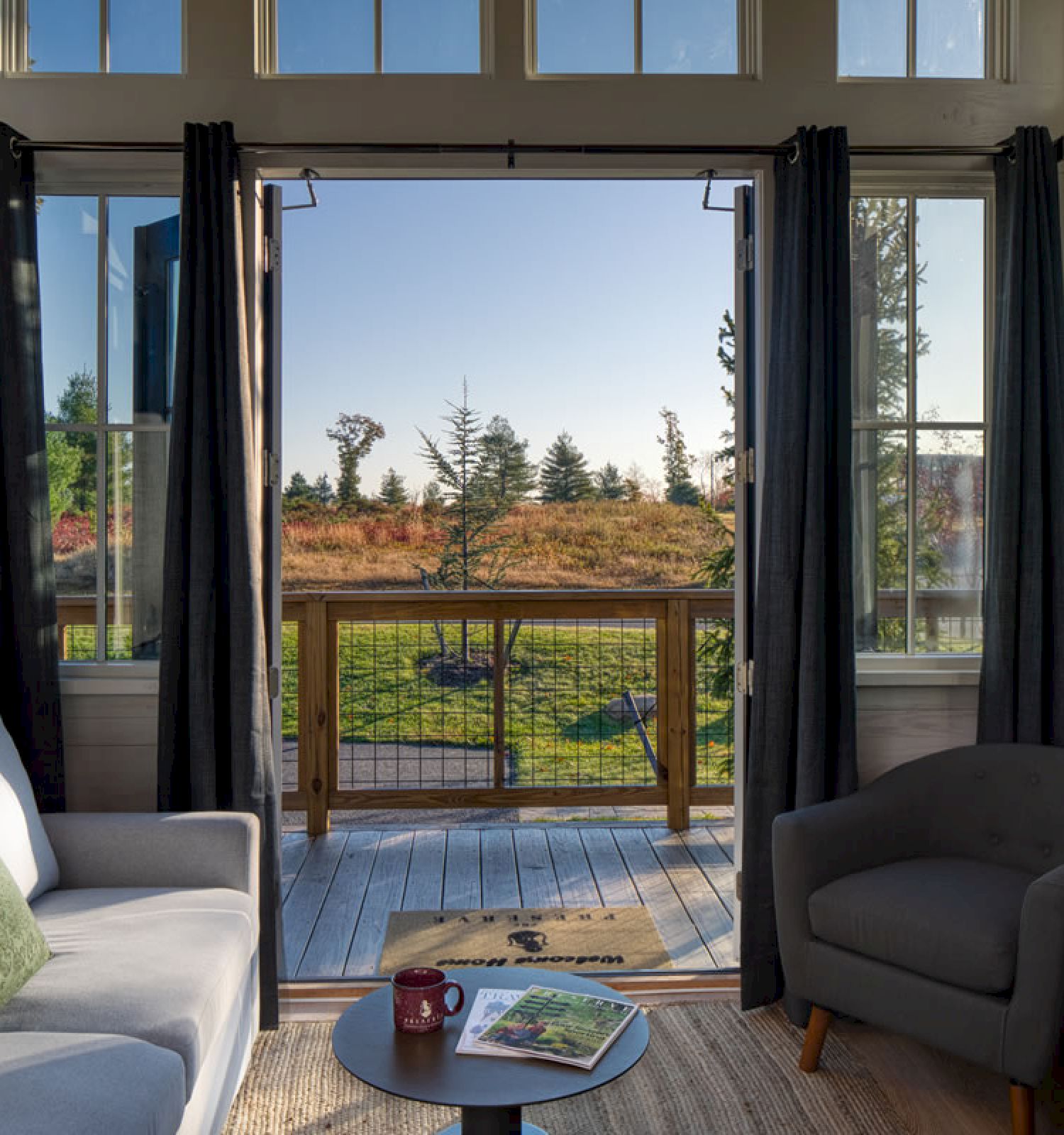 A cozy living room with a gray sofa, armchair, lamp, and coffee table, opening to a wooden deck with a scenic outdoor view and greenery.