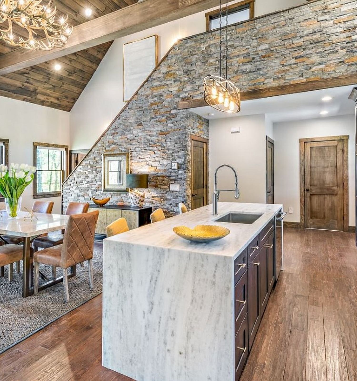 A modern kitchen and dining area with a wooden table, leather chairs, marble island, stainless steel appliances, and a stone accent wall.