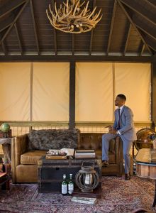 A man in a suit stands in a rustic living room with a leather sofa, antler chandelier, globe, and various decorations, including two bottles on the floor.