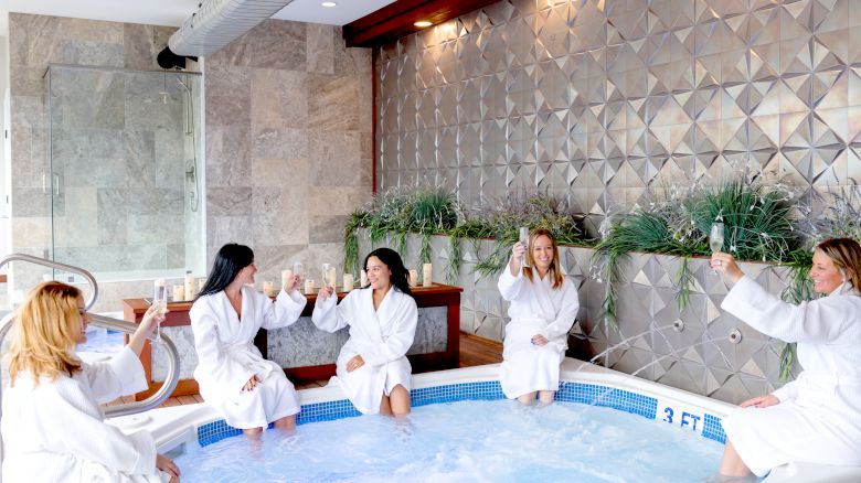 Five people in white robes sitting by a hot tub, raising glasses for a toast in a spa setting with plants and modern design elements.