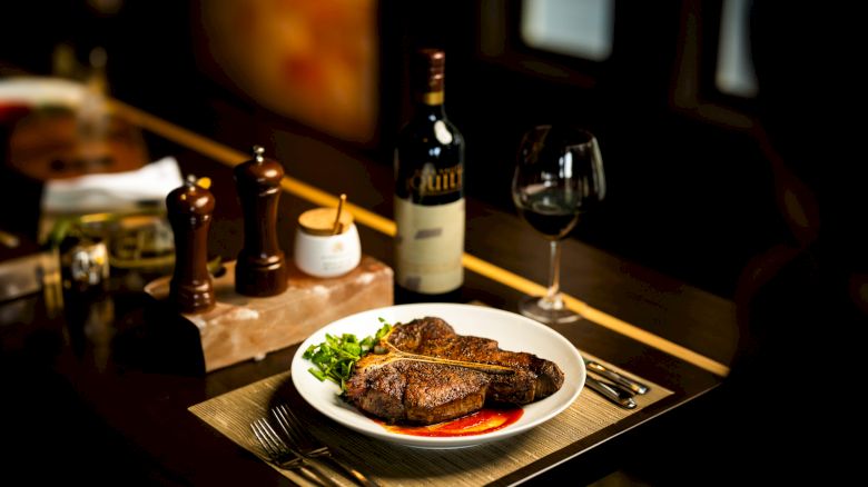 A plate of steak with greens, a bottle of wine, and a glass. Salt and pepper shakers, utensils, and a candle are also set on the table.