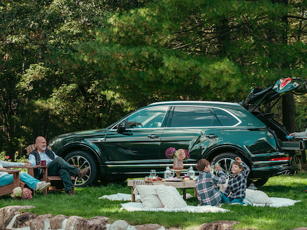 A family enjoys a picnic by an SUV in a lush, green setting with trees, blankets, and food. The SUV's trunk is open for convenience.