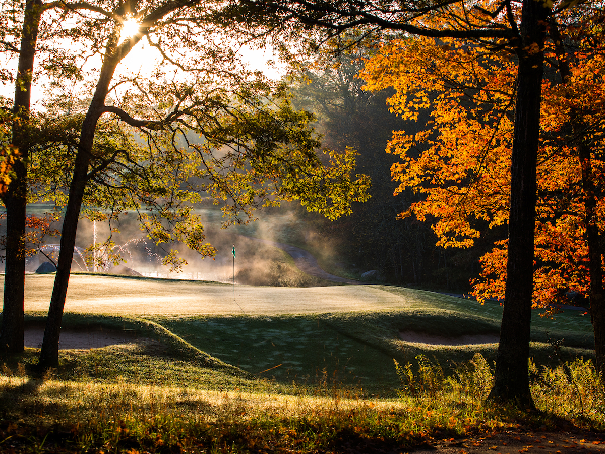 A golf course with morning mist, surrounded by trees displaying fall foliage under a sunrise.