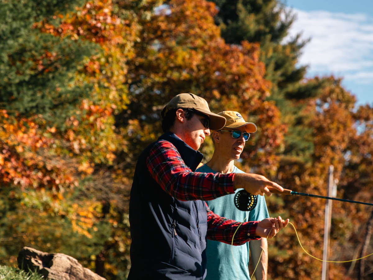 Two people in casual clothes and hats are fly fishing in a scenic outdoor setting with colorful autumn foliage.