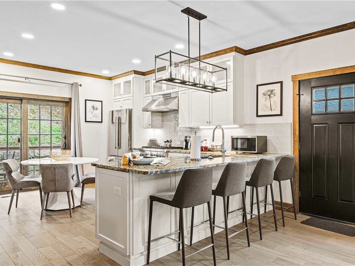 A modern kitchen with an island, bar stools, dining table, and large windows, featuring stylish lighting and decor.