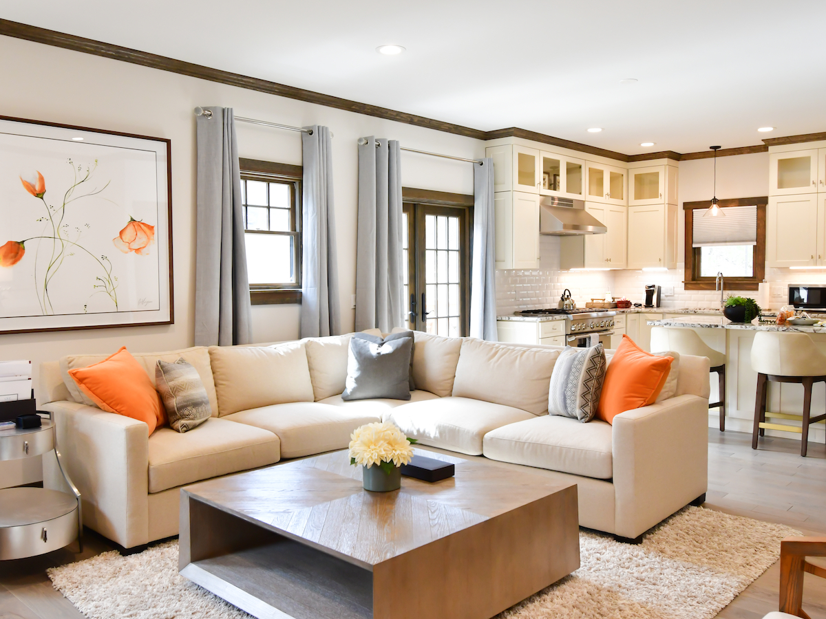 A modern living room and kitchen with a beige sectional sofa, orange cushions, a wooden coffee table, and a small dining area in the background.