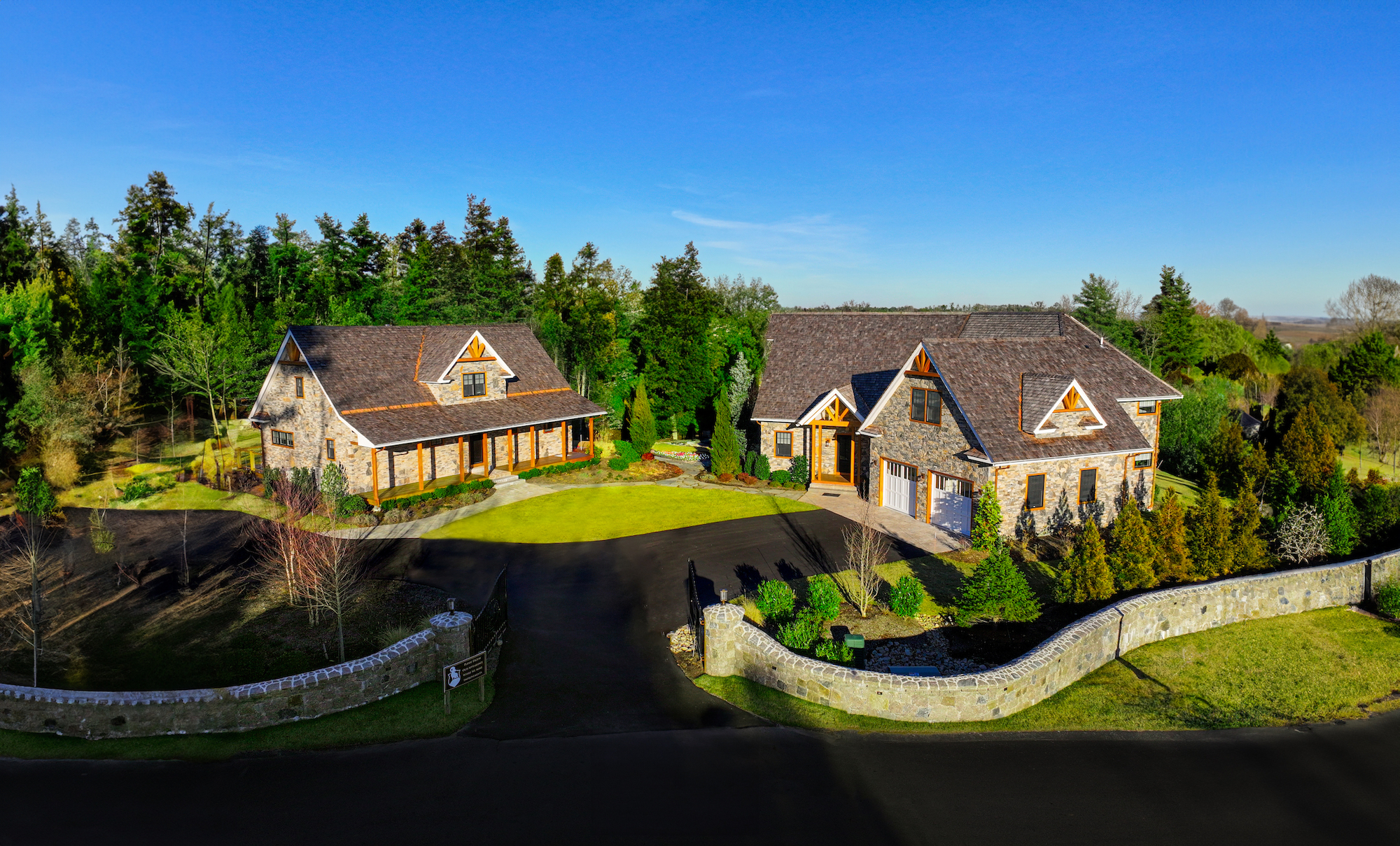 The image shows two large houses with tiled roofs and stone facades, surrounded by landscaped gardens and trees, enclosed by a stone wall.