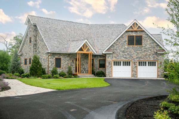 A stone cottage with two white garage doors, a large arched entrance, and a paved driveway surrounded by greenery is pictured.