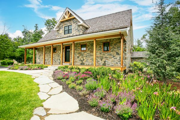 A stone house with a covered porch, surrounded by a well-maintained garden and a flagstone pathway, set against a backdrop of trees and blue sky.