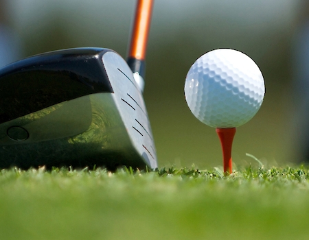 A close-up image of a golf club about to strike a golf ball on a red tee, with grass and blurred background.