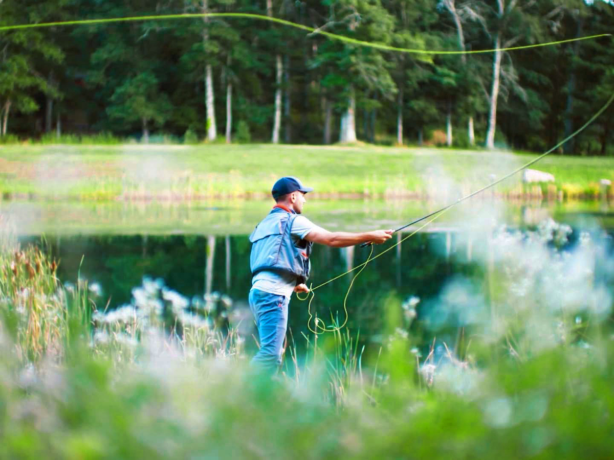 A person is fly fishing near a calm lake, surrounded by greenery and trees, with a focus on casting the fishing line.