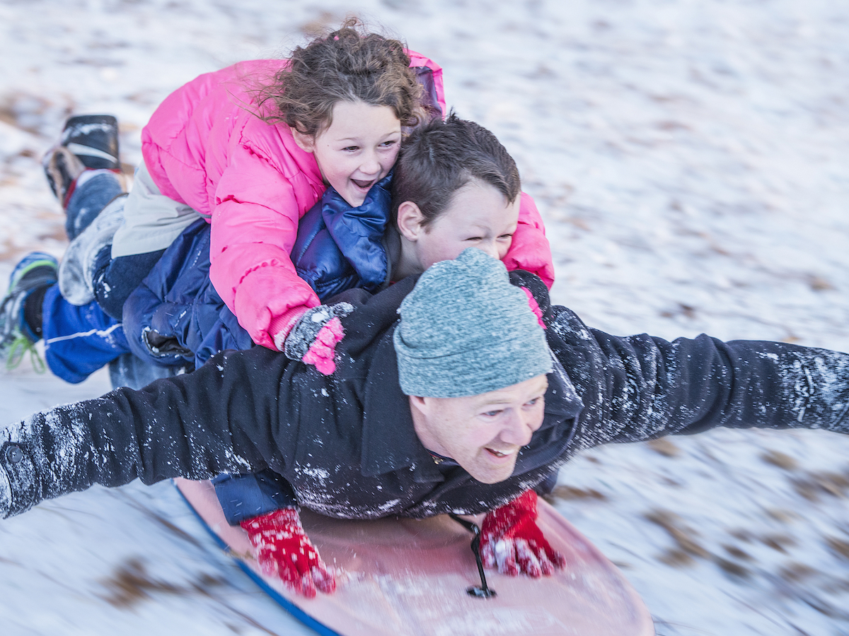 Three people are joyfully sledding down a snowy hill, with two children on top of an adult. The group is having a great time.