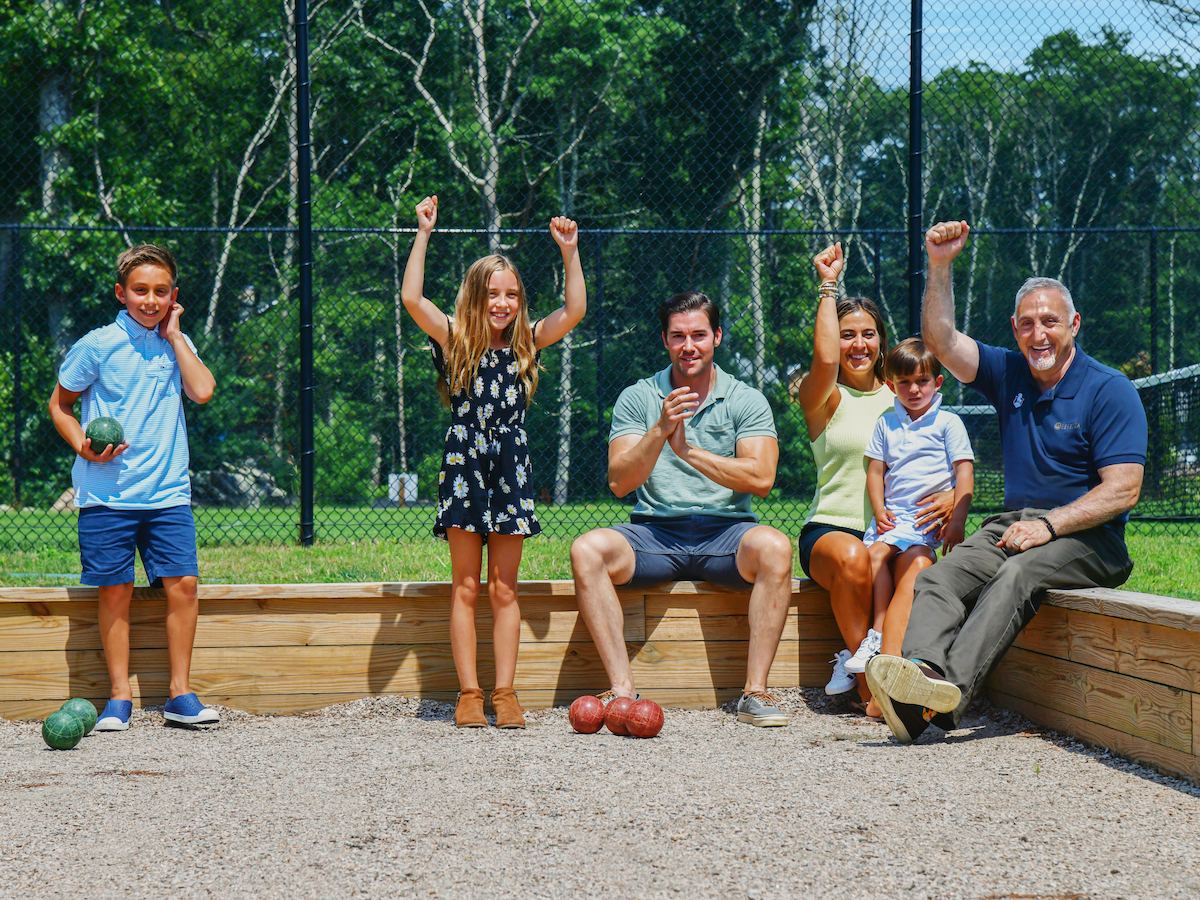 A group of people, including children, celebrate while sitting and standing in a bocce court on a sunny day.