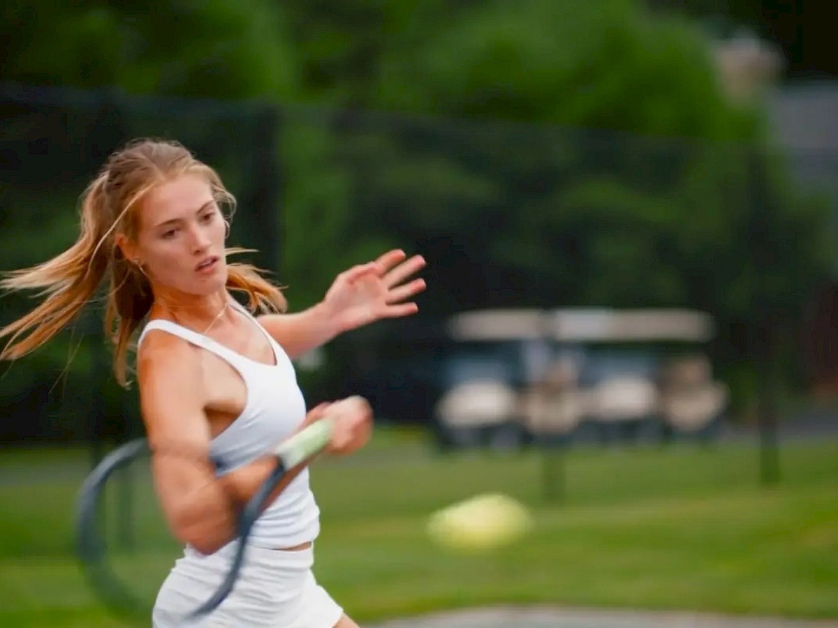 A person playing tennis, captured mid-swing on a grassy court, with a blurred background of greenery and a golf cart.