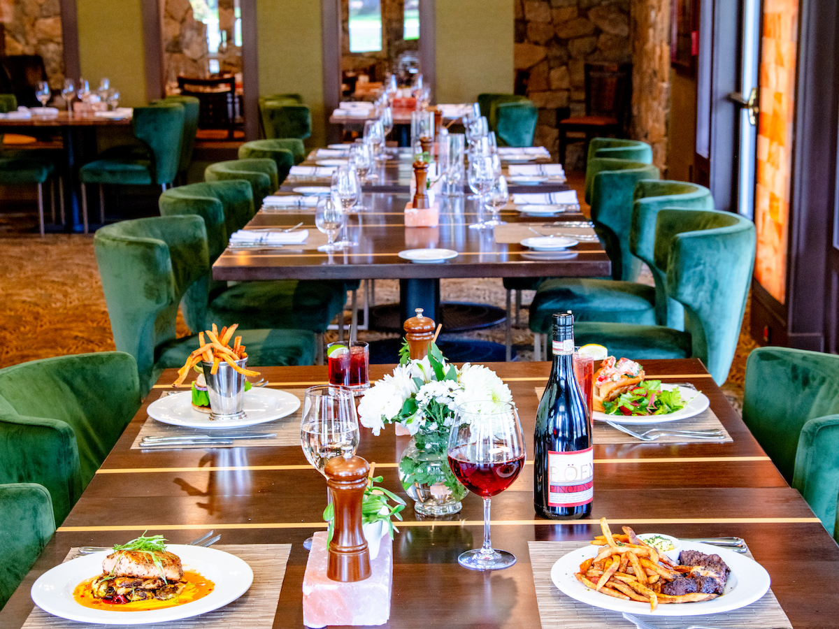 The image shows a dining area with a long table set for a meal, featuring plates of food, wine glasses, and green chairs.