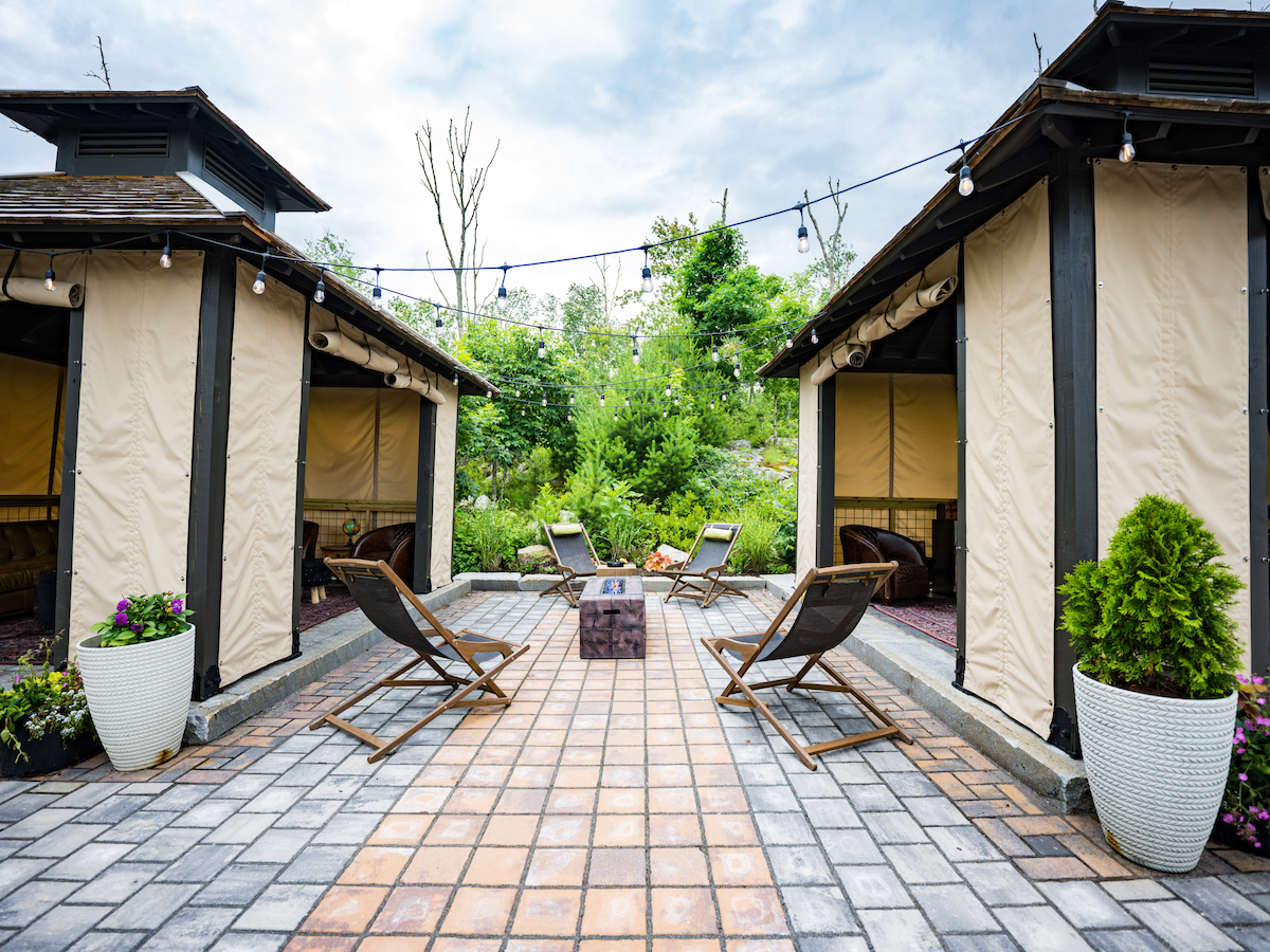 The image shows an outdoor seating area with deck chairs, cabanas, potted plants, string lights, and a central fire pit on a tiled patio.