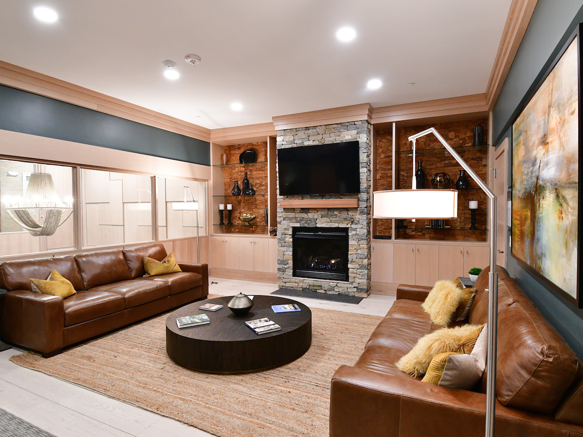 The image shows a modern living room with brown leather sofas, a stone fireplace, wall art, and a large TV, all under bright lighting.