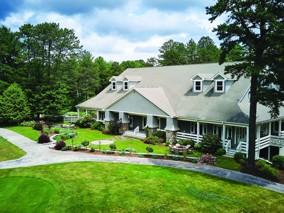 A large house with a sloped roof sits amidst lush greenery and a manicured lawn, surrounded by trees under a partly cloudy sky.