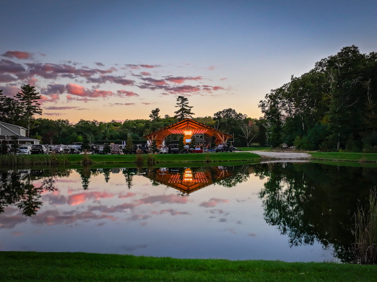 A serene lakeside scene at dusk, featuring a lit wooden structure reflected in the water, surrounded by trees and a colorful sky.