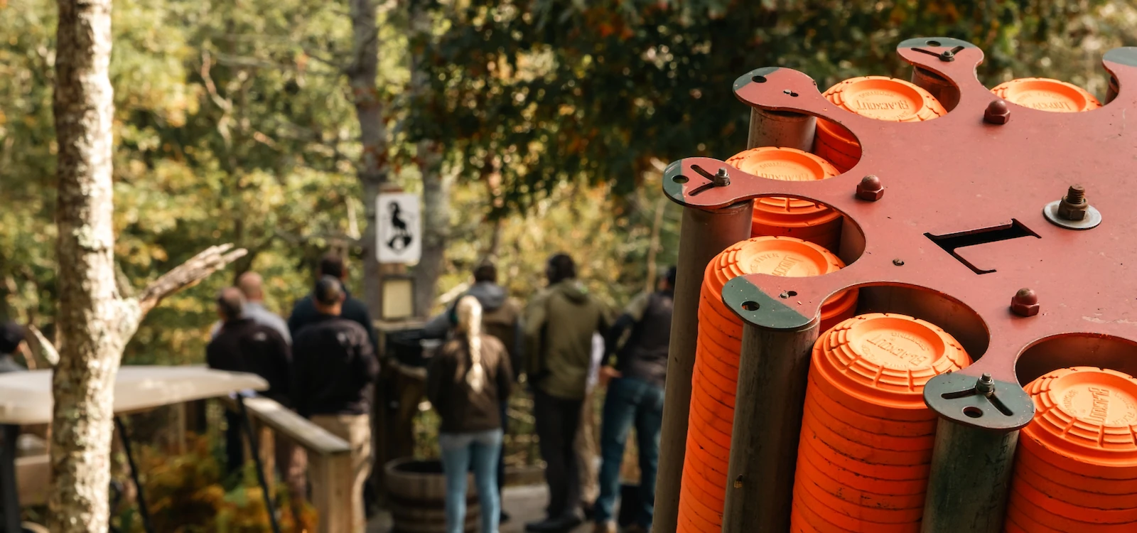 The image shows a group of people outdoors, with a clay pigeon shooting machine loaded with orange disc targets in the foreground.