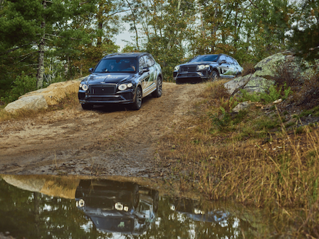 Two SUVs are driving off-road on a dirt path, surrounded by trees and a small water puddle in the foreground, ending the sentence.