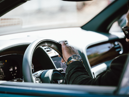 A person wearing sunglasses is driving a luxury car with a prominent wristwatch visible, capturing the interior and dashboard details.