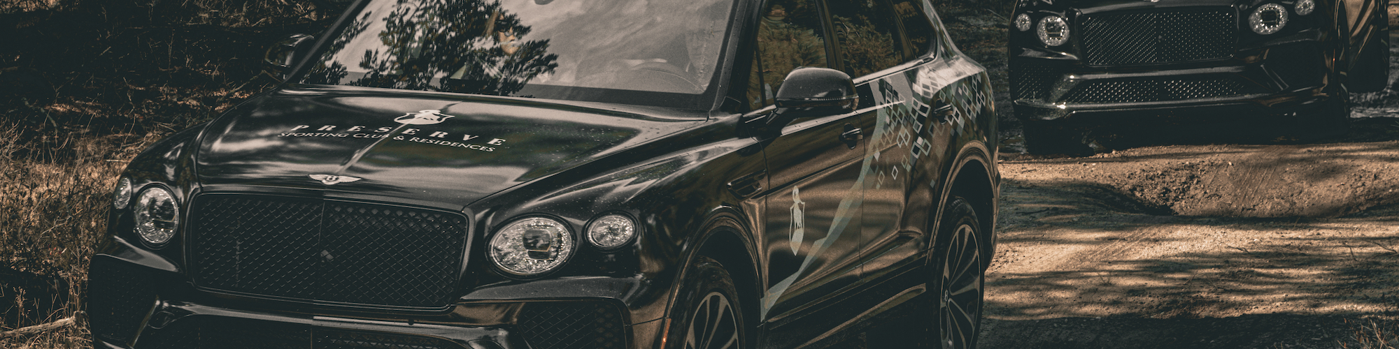 Two luxury SUVs are driving on a dirt road through a forest, showcasing their off-road capabilities.