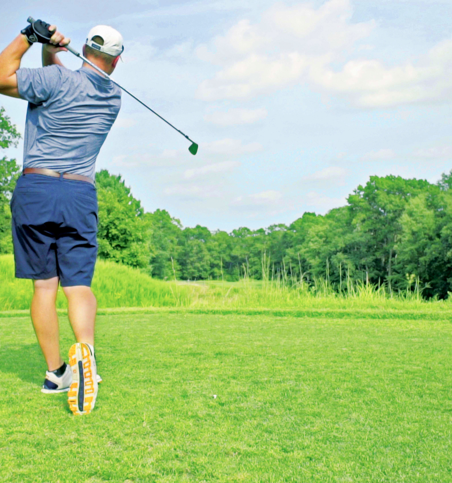 A person is playing golf, swinging a club on a lush, green course with trees and a clear sky in the background.