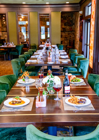 The image shows a long dining table in a restaurant, set with plates of food, wine glasses, and bottles, surrounded by green velvet chairs.